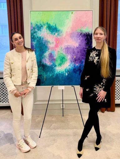 Zwei Frauen stehen neben einem farbenfrohen Gemälde in einem hellen Raum.